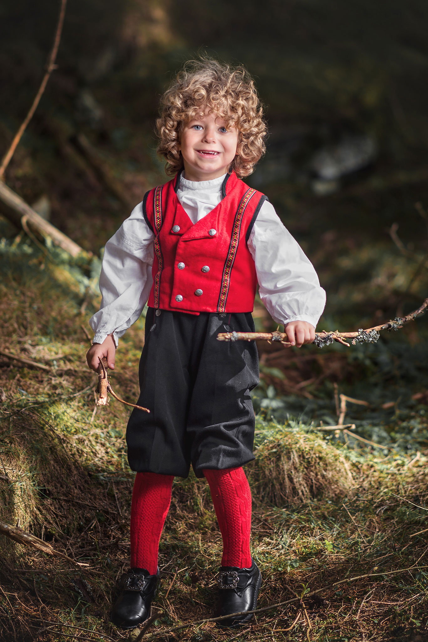 en liten gutt i bunad står og smiler i skogen