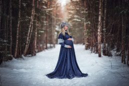 en gravid kvinne i blå kjole står i vinterskogen og poserer til gravidbilder