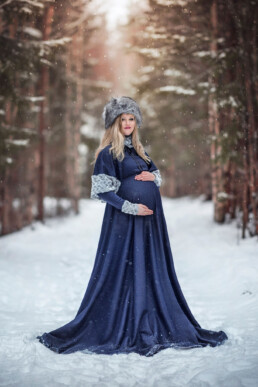 en gravid kvinne i blå kjole står i vinterskogen og poserer for bilder